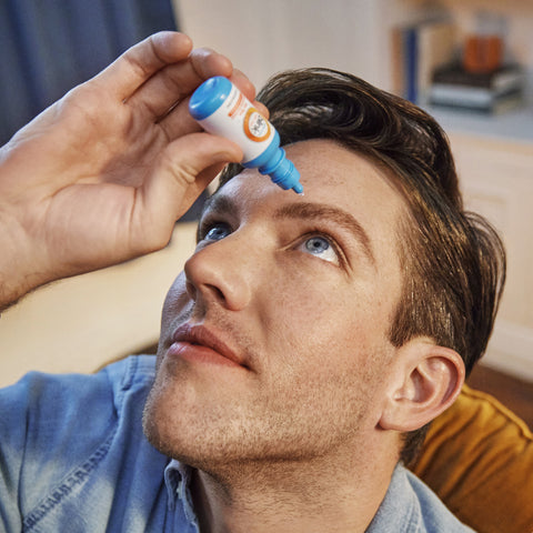 Wearing a denim shirt, a person tilts their head back to use Bausch & Lombs Blink Triple Care 3 in 1 Eye drops with moisture seal technology, soothing dry eyes. The softly blurred background reveals part of a room with a bookshelf.