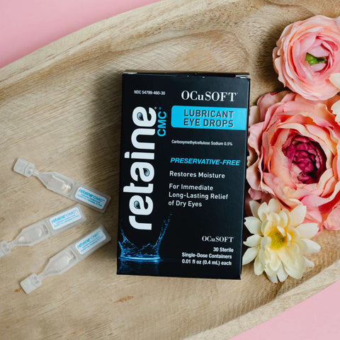 A box of OCuSOFT Retaine CMC Eye Drops for hydrating dry eye relief, featuring preservative-free Carboxymethylcellulose Sodium, rests on a wooden tray surrounded by pink and white flowers, with 30 single-use vials and sleek black packaging.