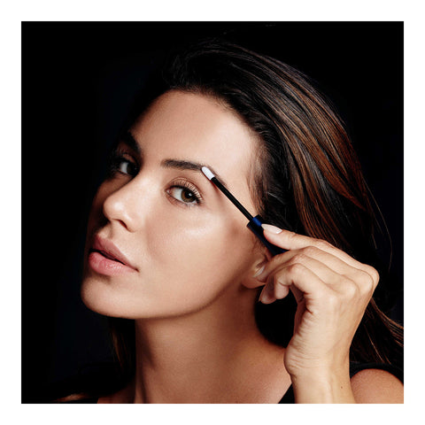 In a dimly lit room, a woman with long dark hair focuses intently as she applies eyebrow mascara using a small brush. The darkness accentuates her features while she subtly incorporates RevitaLashs RevitaBrow Advanced Eyebrow Conditioner Serum for enhanced care and vitality.