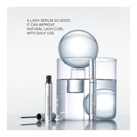 The RevitaLash Advanced PRO Eyelash Conditioner (3.5mL, 6 Mo. Supply) is showcased in a silver tube with an applicator brush before a glass of water; larger glass and spherical objects are in the background. The text highlights its lash-enhancing formula that improves natural curl and conditions brittle lashes daily.