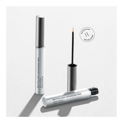 The RevitaLash Advanced PRO Eyelash Conditioner and Enhancing Serum packaging is shown on a light gray surface, with a closed tube beside an open one displaying its fine brush applicator. A circular top-right label reads Physician Developed.