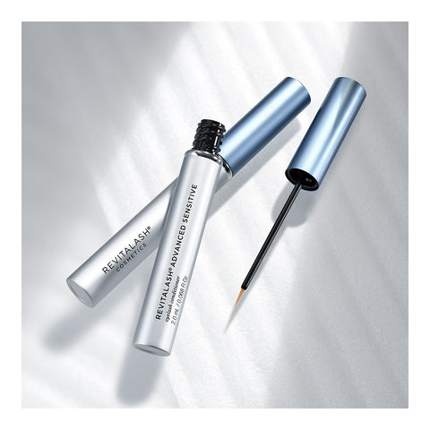 Two metallic blue tubes of RevitaLash Advanced Eyelash Conditioner and Enhancing Serum Sensitive (2.0mL 3 Mo.Supply) are shown; one open with a thin applicator wand, the other closed. Enriched with peptides and botanicals, this duo from RevitaLash rests elegantly on a soft, light grey surface.