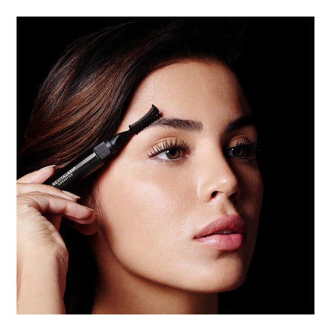 A woman enhances her lashes with precision using RevitaLashs Hi-Def Brow Gel. Her long hair cascades around her face as she focuses intently, showcased against a black background that highlights the meticulous application process.