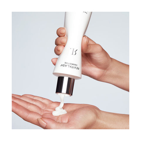 Someone squeezes a creamy white lotion from a RevitaLash tube marked Thickening Conditioner Scalp Therapy Formula, Hypoallergenic (250mL) onto their palm. The shiny silver cap stands out against a simple, light background.