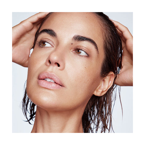 With their hands on their head and a neutral expression, a person with freshly treated wet hair showcases the effects of RevitaLashs Thickening Conditioner Scalp Therapy Formula. Their smooth skin and natural makeup emphasize features like full lips and defined eyebrows against a plain backdrop.