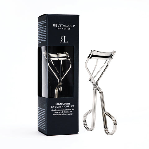 A sleek silver eyelash curler, made from durable carbon steel, is elegantly displayed alongside its black rectangular box. The packaging features RevitaLash and RevitaLash Signature Eyelash Curler in crisp white text.