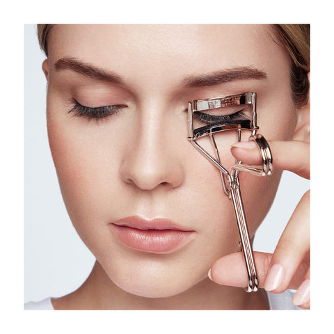 Close-up of someone using a RevitaLash Signature Eyelash Curler on their left eye, displaying a neutral expression with light makeup. Silicone pads gently press the lashes against a plain white background.