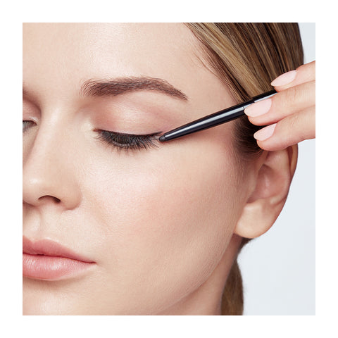 A person uses RevitaLash Defining Liner Eyeliner on their upper eyelid with closed eyes, displaying natural makeup and neutral eyeshadow. In their right hand, the precision of this smudge-proof, high-impact color eyeliner from RevitaLash is evident as BLACK appears in the bottom right corner.