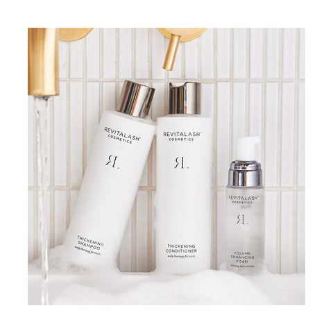 Displayed in a serene bathtub scene with bubbles and running water, the RevitaLash Volumizing Hair Collection Bundle features sleek white bottles with silver caps, including Shampoo, Conditioner, and Foam.