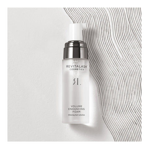 A 55mL bottle of RevitaLash Volume Enhancing Foam for Hair elegantly sits on a textured white surface. The frosted bottle with a silver pump and cap features minimalistic black branding. Enriched with BioPeptin Complex, it complements the wavy design beneath, ideal for thinning hair.