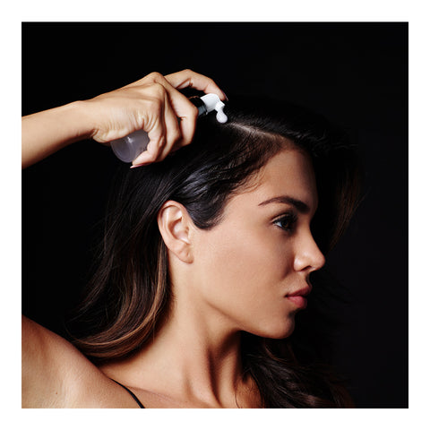 A person with long dark hair sprays RevitaLash Volume Enhancing Foam for Hair (55mL) onto their scalp, the black background highlighting their profile as they apply this BioPeptin Complex-infused solution for thinning hair from RevitaLash.