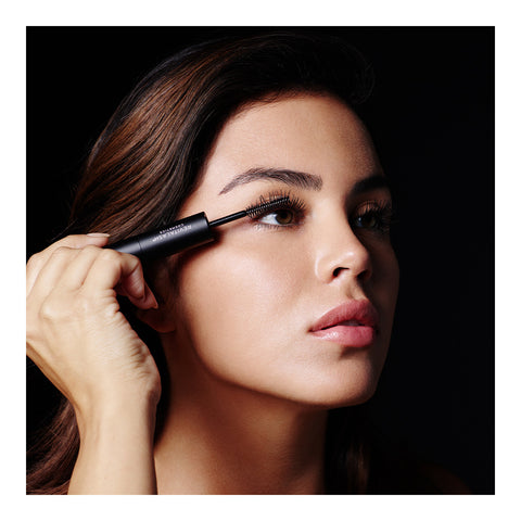 A person with long dark hair uses RevitaLash Double-Ended 2-in-1 Lash Primer & Mascara Volume Set on their upper lashes. The black background accentuates their strong eye makeup and smooth skin, showcasing the transformative power of RevitaLash.