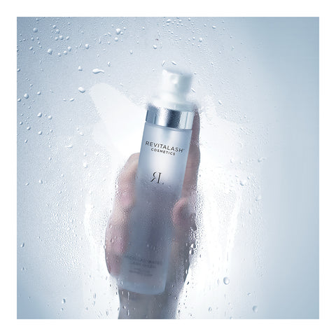 A hand holds a silver and white bottle of RevitaLash Micellar Water Lash Wash, a hypoallergenic makeup remover, suitable for sensitive eyes, against a foggy glass surface. The soft focus creates a fresh, dewy effect.