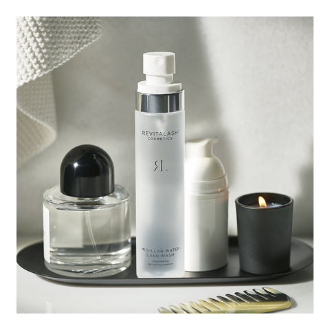 A display on a tray features RevitaLash Micellar Water Lash Wash (100mL), a perfume bottle, a white pump bottle, and a lit candle in a black holder. A gold comb completes the look with a textured towel draped nearby. Hypoallergenic indulgence perfect for sensitive eyes.