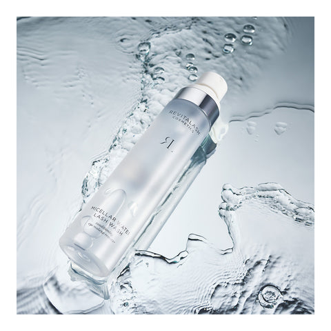 A frosted bottle of RevitaLash Micellar Water Lash Wash, Hypoallergenic Makeup Remover (100mL), sits on a textured wet surface. Ripples and reflections imply motion, while its sleek design with silver accents and clear labeling showcases the formula, safe for sensitive eyes.