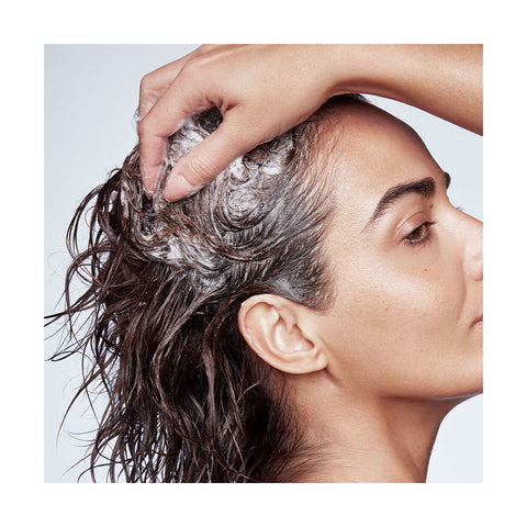 A person with wet hair uses RevitaLash Thickening Shampoo, massaging it onto their scalp. The side profile of their face is visible as they work the foam for effective scalp therapy, set against a plain, light-colored background that accentuates the soothing ritual.