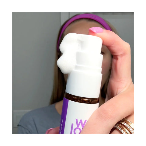 A person with pink nails presses the pump on a white and purple bottle of We Love Eyes SuperGLA eyelid & eyelash foaming cleanser, releasing non-toxic white foam. They wear a pink headband, with their face mostly obscured in the background.