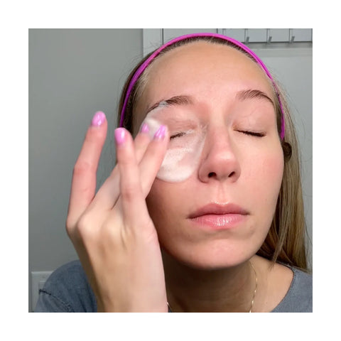 Wearing a pink headband and gray top, a person with long hair and purple nails applies the We Love Eyes SuperGLA eyelid & eyelash foaming cleanser to their face using their fingers, enjoying its non-toxic formula as they keep their eyes closed in this tranquil skincare ritual.